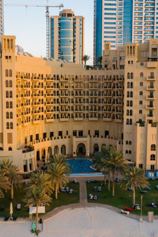 Aerial view of a luxury hotel in Ajman city surrounded by palm trees and urban landscape.